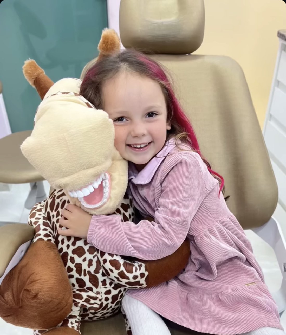Pacientinha sorrindo na cadeira do consultório, abraçada a uma girafa de pelúcia